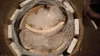 The image shows a product inside a cardboard box, wrapped in plastic and protective foam. It appears to be a water heater or boiler. Produktbild-Vorschau