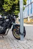 A black Yamaha motorcycle is parked on a cobblestone street, locked to a pole with a chain lock. A modern building and trees are in the background. Anteprima dell’immagine prodotto