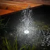 A solar-powered fountain with LED lights is operating in a pond next to a wooden deck. Produktbild-Vorschau