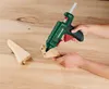A person is using a green hot glue gun to attach two pieces of wood together on a wooden surface. Produktbild-Vorschau