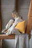A woman sits on a wooden beam, holding a yellow cushion and a gray blanket. Produktbild-Vorschau