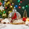 A festive Christmas scene with a glass ornament containing a red cardinal, surrounded by snow, pinecones, candles, and a Christmas tree with lights. Produktbild-Vorschau