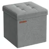 A gray fabric storage ottoman with a tufted lid and a brown logo tag on the side, displayed against a white background. Produktbild-Vorschau
