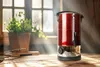 A red beverage dispenser sits on a wooden table in front of a window with sunlight streaming through. Náhled obrázku produktu