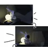 A rabbit-shaped night light projector sits on a bedside table next to a book, casting a warm glow. Náhled obrázku produktu