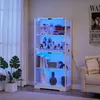 A white glass display cabinet with LED lighting, filled with various collectibles and decorative items, is shown in a room setting next to a sofa and a large plant. Náhled obrázku produktu