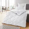 A bedroom scene with a neatly made bed featuring a white comforter and pillows, a chair with a blanket, and a small side table with decorative items. Produktbild-Vorschau