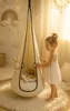A young girl in a white dress stands next to a hanging pod chair in a child's room, with a book and stuffed animals inside. Produktbild-Vorschau