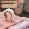 A roll of white hemming tape sits on top of folded pink and purple fabric, with text overlays indicating product features. Náhled obrázku produktu