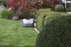 A robotic lawnmower is shown on a green lawn next to a manicured hedge, with trees and rocks in the background. Produktbild-Vorschau