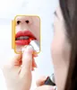 A woman is applying red lipstick while looking in a small, yellow, rectangular mirror held in her hand. The mirror reflects her lips and the lipstick. Voir les images du produit