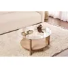 A round coffee table with a glass top and wooden base is placed on a fluffy rug next to a sofa. Produktbild-Vorschau
