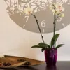 An orchid plant in a purple pot sits on a dark surface in front of a wall decorated with a large clock face graphic. Two decorative palm leaves filled with small items are in the foreground. Produktbild-Vorschau