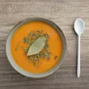 A bowl of orange soup garnished with herbs and a bay leaf, next to a white spoon on a wooden surface. Anteprima dell’immagine prodotto