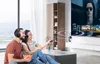 A couple is sitting on a couch watching TV and listening to audio through headphones. The man is holding a bowl of snacks, and the woman is holding a remote control. Produktbild-Vorschau