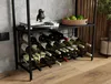A metal wine rack filled with wine bottles, placed on a wooden floor next to a concrete wall. There are also some books and a decorative bird on the shelf above the wine rack, and a basket of apples. Podgląd zdjęcia produktu