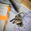 A close-up of a keychain with a lucky angel charm and clover, placed on top of books and papers with handwritten notes and highlighted text. Produktbild-Vorschau