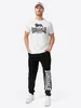 A man is standing and posing for a product shot, wearing a white t-shirt and black sweatpants. Produktbild-Vorschau