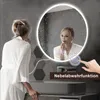 A woman in a white robe is applying makeup in front of a round LED mirror in a bathroom. The mirror has a defogger function indicated by a touch icon and text. Produktbild-Vorschau