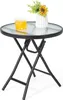 A small round glass-top table with a black metal folding frame, a glass of yellow liquid, and a vase with flowers on top. Produktbild-Vorschau