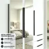 A close-up view of a white wardrobe with mirrored doors, showing black metal handles and drawers. A bedroom interior is reflected in the mirrored doors. Produktbild-Vorschau