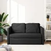 A dark grey sofa bed is shown in a well-lit room with a large plant on the left and a bookshelf on the right. Produktbild-Vorschau