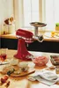 A red KitchenAid stand mixer with a meat grinder attachment is set up on a wooden kitchen counter, surrounded by ingredients like ground meat, onions, and eggs. Náhled obrázku produktu