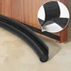 A close-up of a black foam door seal with Velcro attachment, placed on a wooden door and wooden floor. Náhled obrázku produktu