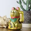 A golden Maneki Neko cat figurine sits on a wooden surface, with a seashell and a potted plant in the background. Produktbild-Vorschau