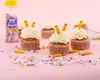 A group of carrot-themed cupcakes decorated with white frosting and small carrot sprinkles, arranged on a pink background with scattered sprinkles. Náhľad obrázku produktu
