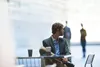 A man in a suit sits at a table with coffee cups, looking away from the camera. Other people are blurred in the background. Podgląd zdjęcia produktu