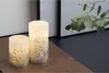 Two LED candles with glitter accents sit on a wooden surface, with a branch of eucalyptus and framed artwork in the background. Produktbild-Vorschau