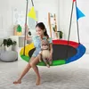 A young girl sits on a colorful nest swing, holding a stuffed deer toy. The swing is in a room with furniture and decorations. Náhled obrázku produktu