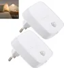 Two white LED night lights with motion sensors are shown against a white background. A smaller inset image shows one of the lights plugged into a wall socket next to a bed. Produktbild-Vorschau