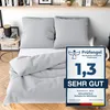 A bed with light gray bedding, including a duvet and pillows, with a product test seal on the right. Produktbild-Vorschau