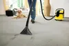 A person is using a carpet cleaner on a carpeted floor, with a dog lying down in the background. Náhled obrázku produktu