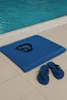 A blue towel with swimming goggles and flip-flops next to a swimming pool. Produktbild-Vorschau