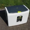 A white and gray cooler box with a green handle is placed on a paved surface, with grass in the background. Produktbild-Vorschau