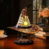 A witch hat-shaped stained glass lamp sits on a wooden table next to a cup of coffee and a stack of books, with flowers in the background. Produktbild-Vorschau