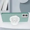 A green phone case with a clear PopGrip attached is shown on a white surface, with a blurred background of books and a wall. Produktbild-Vorschau
