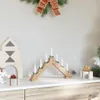 A wooden candle bridge with seven white LED candles, decorated with small green sprigs, sits on a white surface next to a wooden house-shaped decoration and wrapped gifts. A Christmas wreath with a red and green bow hangs above. Voir les images du produit