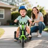 A child is riding a green tricycle outdoors, with a woman crouching behind them. Produktbild-Vorschau