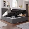 A bedroom scene with a bed that has its storage compartment open, revealing bedding and pillows. The room has artwork on the wall and a window with sheer curtains. Produktbild-Vorschau