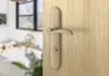 A close-up of a bronze door handle on a wooden door, with a blurred bathroom in the background. Produktbild-Vorschau