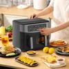 A person's hand is shown operating a dual-basket air fryer on a wooden countertop, with various food items like chicken, fries, fruit slices, and pastries arranged around it. Produktbild-Vorschau