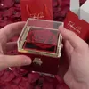 Hands holding a red rose in a clear and red gift box, surrounded by red rose petals. A red gift bag and box are in the background. Anteprima dell’immagine prodotto