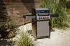 A gas grill is placed on a gravel patio next to tall green grass and a wooden fence. Produktbild-Vorschau