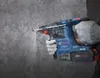 A hand wearing a glove is holding a blue and black Bosch rotary hammer drill and drilling into a gray concrete wall. Voir les images du produit