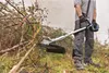 A person is using a cordless string trimmer to clear overgrown brush and branches. Voir les images du produit