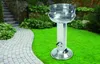 A stainless steel charcoal grill on a pedestal in a garden setting with a paved walkway and stone steps. Produktbild-Vorschau